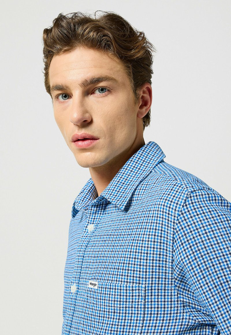 Camicia a quadratini blu e bianchi con colletto classico, chiusura a bottoni e un piccolo etichetta con il marchio sulla tasca del petto. Tessuto dalla superficie liscia.