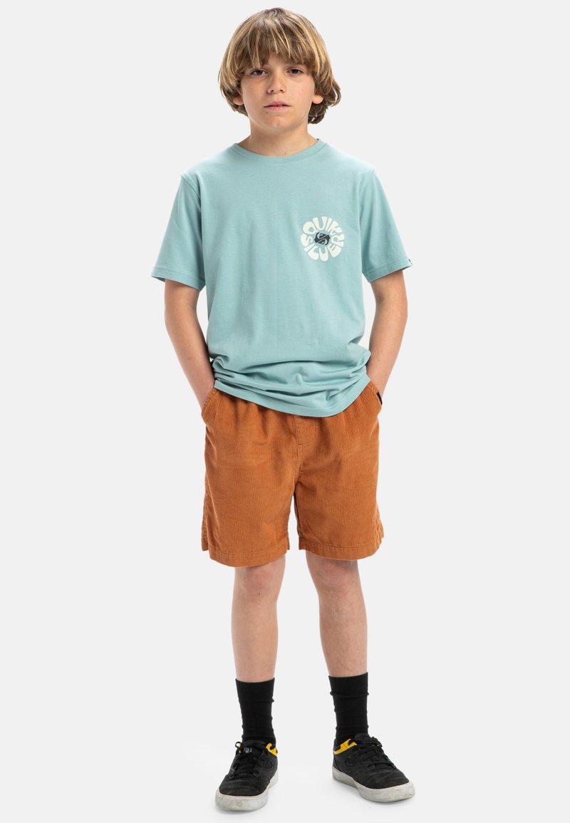 Camiseta de algodón azul claro con un logotipo blanco circular en el pecho, combinada con pantalones cortos de pana naranja. Calcetines negros y zapatillas deportivas oscuras completan el conjunto.