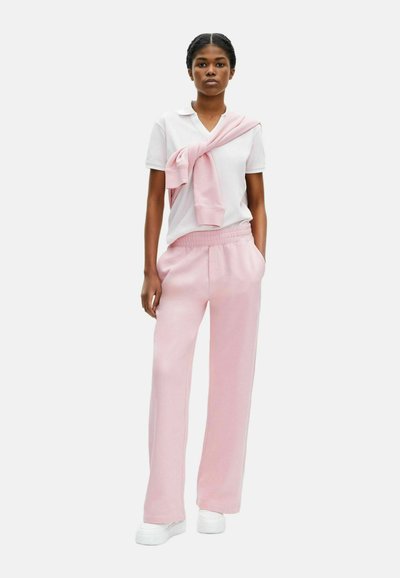 Roze sweatpants met een ontspannen pasvorm en een elastische tailleband, gecombineerd met een wit polo-shirt. Lichtroze trui over de schouders gedrapeerd. Witte sneakers.