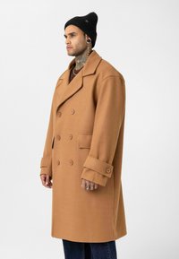 Man met nek- en handtattoos, gekleed in een beige dubbelgeknoopt jas, zwarte muts en donkere spijkerbroek, staand tegen een witte achtergrond.