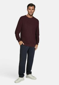 Bordeauxroter Strickpullover mit rundem Halsauschnitt, lockerer Passform und langen Ärmeln, kombiniert mit dunklen Hosen und weißenSneakern vor einem neutralen Hintergrund.