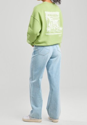 Personne portant un sweat-shirt vert clair avec un graphisme de devanture de magasin blanche dans le dos, un jean bleu clair et des baskets blanches, debout devant un fond uni.