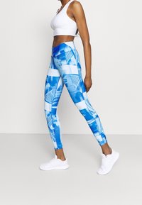 Modré legíny s potiskem mají abstraktní vzory v různých odstínech modré, spárované s bílým crop topem a bílými sportovními botami.