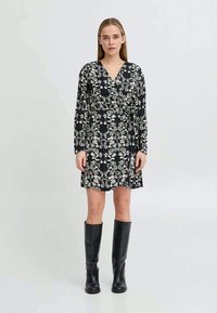 Ženska v črni obleki z zavojem in belim cvetličnim potiskom ter dolgimi rokavi, ki jo dopolnjujejo črne škornje do kolen, stoji ob enostavni ozadju.