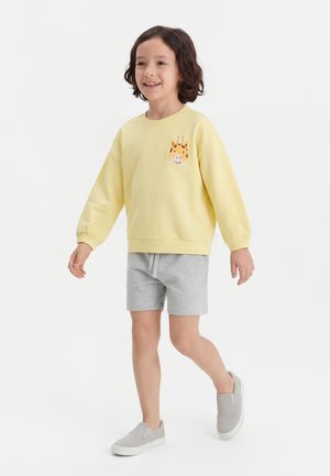 Barn der går iført en gul sweatshirt med et girafmærke, grå shorts og grå slip-on sko på en hvid baggrund.