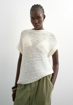 Kvinde med flettet hår iført en løs hvid strikket top og olivengrønne bukser, stående med den ene hånd i lommen mod en ensfarvet baggrund.