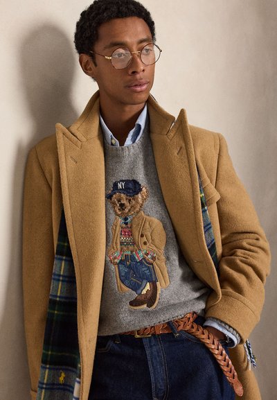 Jeune homme portant des lunettes rondes, un manteau camel sur un pull gris avec un motif d’ours en peluche, une chemise bleue, un jean et une ceinture tressée.