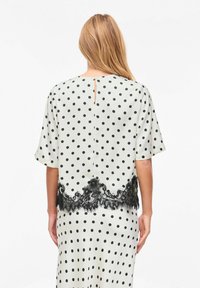 Blouse à pois crème, avec manches courtes, fermeture au dos en trou de serrure, et bordure en dentelle noire à l'ourlet pour un détail supplémentaire.