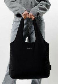 Svart tote-väska i mjukt material, med en rundad topp och breda, platta handtag. Diskret logotyp på framsidan, rektangulär form.