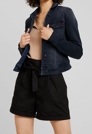 Persona che indossa una giacca di jeans scura sopra un top beige a coste e pantaloncini neri a vita alta con cintura di tessuto annodata.