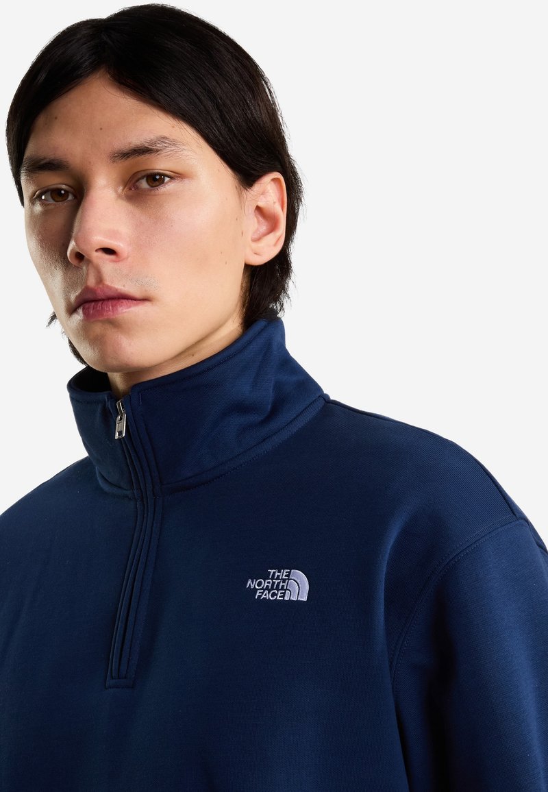 Joven con cabello oscuro de longitud media lleva un polar The North Face azul marino con cremallera hasta el pecho sobre un fondo claro y liso.