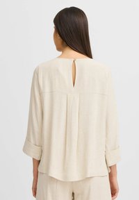 Haut en lin beige avec une ouverture en forme de clé à l'arrière, manches longues retroussées aux poignets et coupe décontractée. Tissu texturé avec des nuances subtiles.