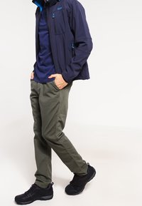 Veste softshell bleu marine avec accents bleus, pantalon kaki et chaussures de randonnée noires. La veste est dotée de poches zippées et d'une finition texturée.