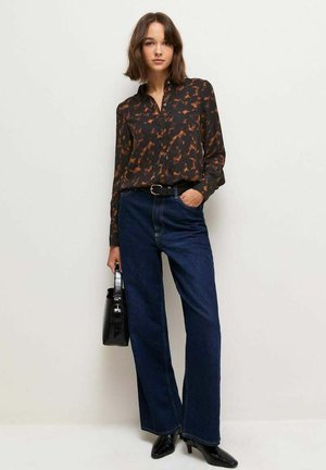 Jeune femme portant un chemisier à motifs noirs et marrons, un jean large bleu foncé, des talons noirs, et tenant un sac à main noir.