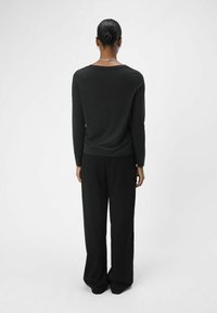 Schwarzes Langarmshirt mit rundem Ausschnitt, kombiniert mit schwarzen weit geschnittenen Hosen, von hinten vor einem hellen Hintergrund gezeigt.