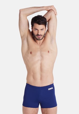 Pantalones de baño azul marino fabricados con un material liso y elástico que presenta un logo "arena" blanco en el lado izquierdo, llevados por un modelo que se está estirando.