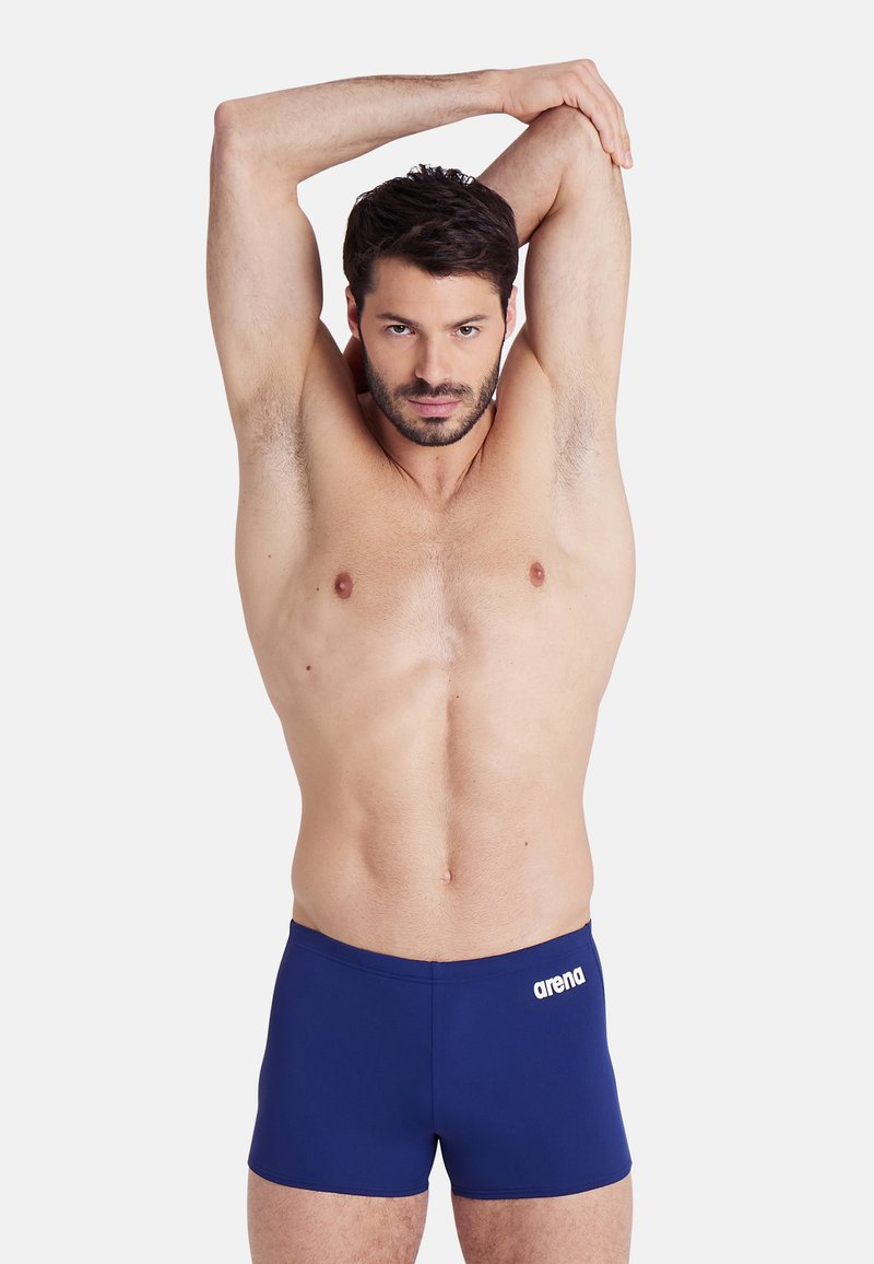 Dunkelblaue Badehose aus glattem, dehnbarem Material mit einem weißen "arena"-Logo auf der linken Seite, getragen von einem Modell beim Dehnen.