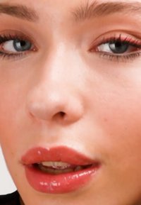 Nahaufnahme eines Gesichts mit heller Haut, blauen Augen und vollen Lippen, die mit glänzendem pinkfarbenem Lippenstift bedeckt sind. Ein dezentes Rouge und weiche Wimpern sind sichtbar.