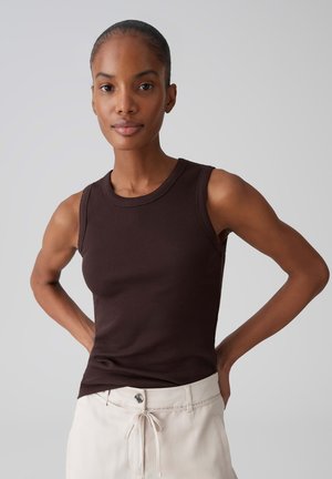 Jeune femme aux cheveux tirés en arrière portant un haut sans manches côtelé marron foncé et un pantalon beige clair, debout les mains sur les hanches.