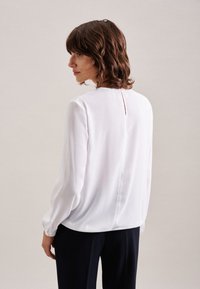 Blusa bianca a maniche lunghe con scollatura tonda e dettagli a chiave nella parte posteriore, realizzata in tessuto liscio con una leggera lucentezza. Abbinata a pantaloni scuri.