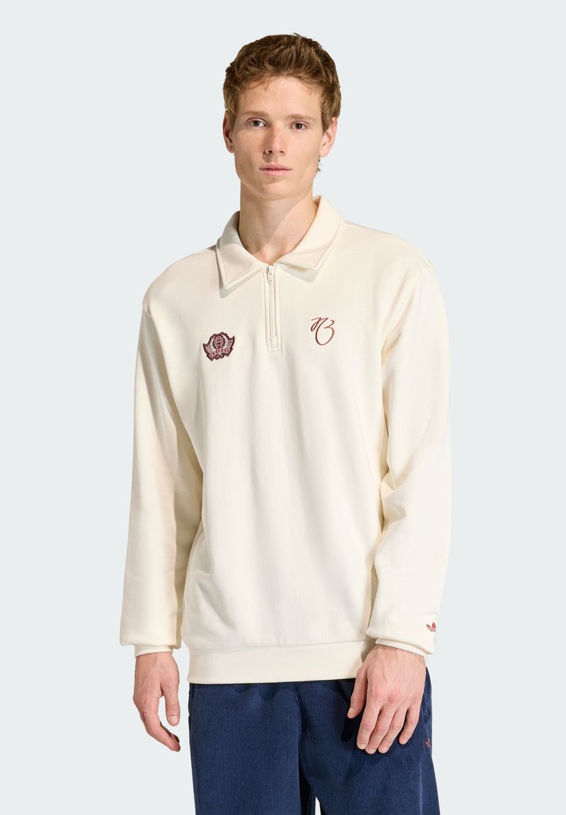 Pull-over couleur crème avec col à fermeture éclair, arborant des logos brodés en bordeaux. Texture douce, manches longues et coupe décontractée.