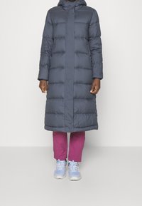 Manteau rembourré gris foncé avec capuche, présentant une coupe droite, un design matelassé et une ligne d'ourlet arrivant au mollet. Associé à un pantalon rose et des baskets.