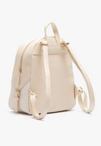 Krémszínű műbőr hátizsák állítható pántokkal, aranyszínű cipzárokkal és szőtt oldalsó panelekkel. Simább textúrájú, kompakt formájú.