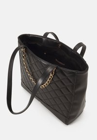 Bolso tote negro acolchado con un acabado de cuero suave, acentos de cadena en tono dorado y dos asas para el hombro. El interior incluye un bolsillo con cremallera.