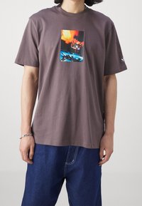 Brun bomull t-shirt med rund hals, prydd med en rektangulär grafisk tryck i livliga färger centrerad på framsidan. Kortärmad.