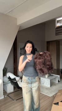 Smeđa prsluk od umjetnog krzna s pahuljastom teksturom, koji drži osoba odjevena u sivu majicu bez rukava i istrošene svijetloplave široke traperice.