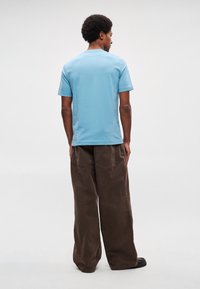 Helehall sinine puuvillane T-särk ümar kaelusega, millele on kombineeritud lahedalt istuvad pruunid sametpüksid ja mustad jalatsid, vaadates tagantpoolt.