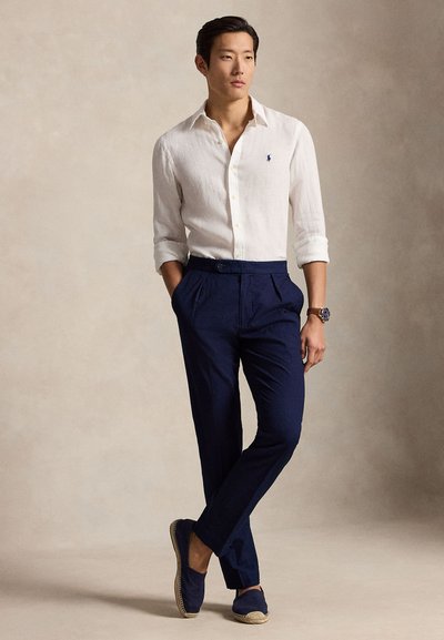 Άνδρας που φοράει λευκό λινό πουκάμισο με κουμπιά, ναυτικό μπλε πλισέ παντελόνι, μπλε εσπαντρίγιες και ρολόι χειρός, στέκεται με τα χέρια στις τσέπες.