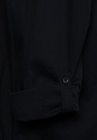 Schwarzer Hemdmanschette mit einem Knopfdetail. Der Stoff wirkt glatt und leicht strukturiert, mit einem klaren, minimalistischen Design.