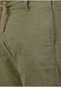 Olive green linen blend pants with a textured surface, drawstring waist, and side pockets. Features a brown leather tag near the waistband.