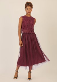 Bordeauxrotes Kleid mit Spitzen-Oberteil und gewellten Kanten sowie einem Tüllrock, der mit Punkten verziert ist. Schwarze Ankle-Strap-Absätze runden das Outfit ab.