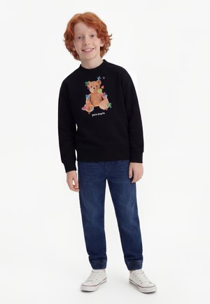 Smiling red-haired boy wearing a black sweatshirt with a teddy bear graphic, blue jeans, and white sneakers, standing against a white background.