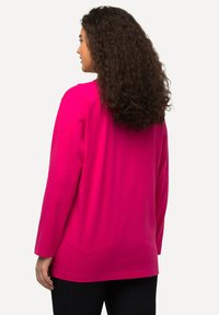 Fuchsiová dlouhá halenka vyrobená z měkké bavlny, s volným střihem a zaobleným lemem, kombinovaná s tmavě modrými džínami.