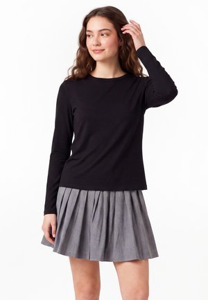 Jonge vrouw met een zwart topje met lange mouwen en een grijze geplooide rok, die haar haar aanraakt en opzij kijkt met een lichte glimlach.