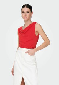 Top sans manches rouge avec un décolleté drapé, associé à une jupe en denim blanche taille haute avec une fente sur le devant et des poches latérales.