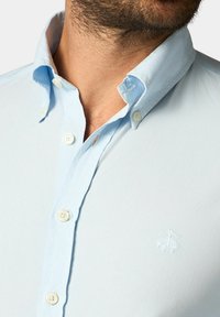 Chemise boutonnée bleu clair avec une texture douce, présentant un col classique, des boutons blancs et un logo brodé sur la poitrine.