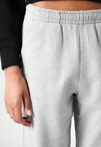 Lichtgrijze joggingbroek met een elastische tailleband, voorzien van zijzakken en een zachte, gestructureerde stof. Hand rustend op het been.