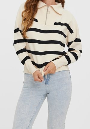 Femme portant un pull crème avec des rayures horizontales noires et un col zippé, associé à un jean bleu clair, mains ajustant l'ourlet du pull.