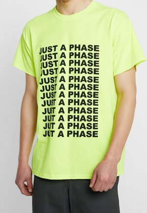Osoba ubrana w jaskrawożółtą koszulkę z powtarzającym się, pogrubionym czarnym napisem "JUST A PHASE" ułożonym w wzór siatki na przodzie.