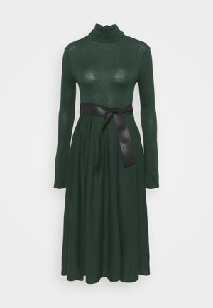 Długa sukienka z golfem w ciemnozielonym kolorze, z dopasowanym gorsetem, zwiewną spódnicą i szerokim czarnym paskiem zawiązanym w talii.