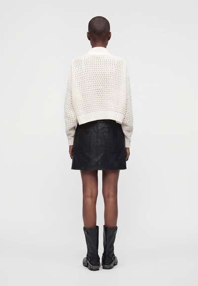 Personne aux cheveux courts portant un pull en maille blanc, une jupe en cuir noire et des bottes noires mi-mollet avec des détails en chaîne, debout de dos.