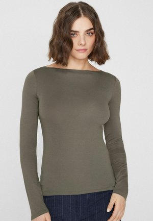 Kobieta o falowanych brązowych włosach do ramion, ubrana w oliwkowo-zieloną dopasowaną bluzkę z długim rękawem i granatowe spodnie na jednolitym tle.