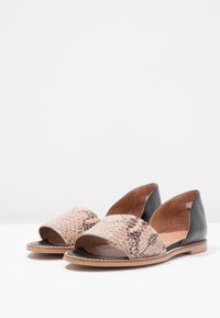 Ett par platta sandaler med framsnören i tan-färgad orm-mönster och svarta läderpaneler på baksidan, placerade på en vit yta.