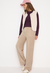 Vrouw in een roze pet, beige fleecevest, paarse trui, beige wijde broek en sneakers die tegen een witte achtergrond staat.
