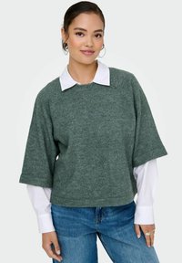 Groene gebreide trui met korte, brede mouwen, geribbelde structuur en een witte overhemd met kraag eronder, gecombineerd met blauwe jeans.
