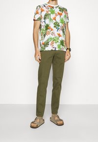 Camisa de manga corta con estampado floral en tonos verde, naranja y blanco; pantalones ajustados en verde oliva; sandalias grises con detalles de hebilla.